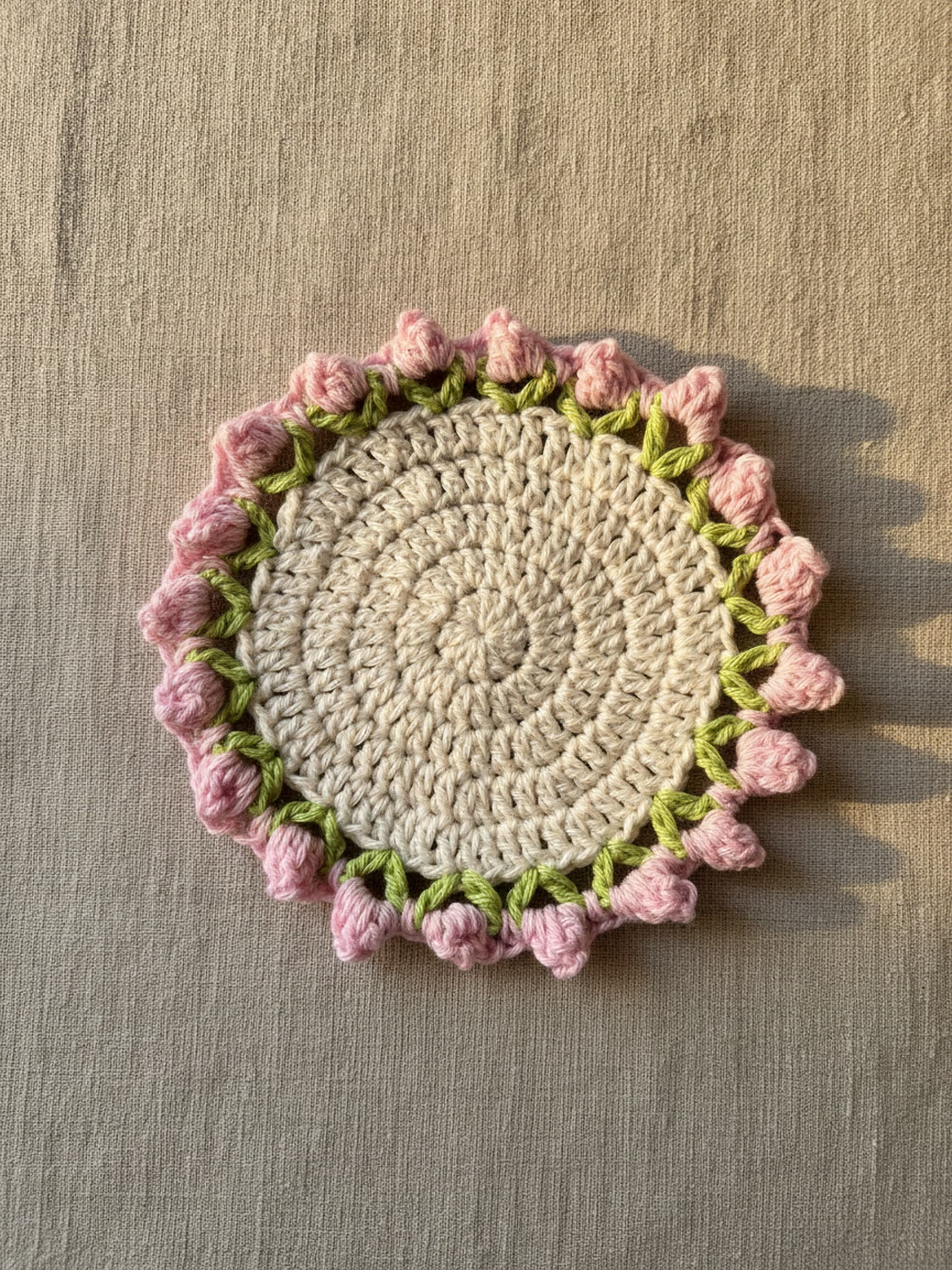 Crochet rosebud coaster laid flat on oatmeal linen, cream centre with pink rosebud and green leaf border